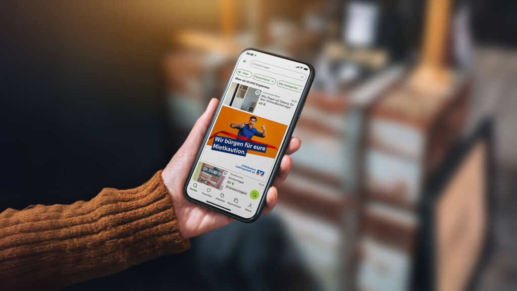 Ein Smartphone in einer Hand. Auf dem Bildschirm ist die App "Kleinanzeigen" geöffnet und eine Anzeige zum Thema Mitkaution.