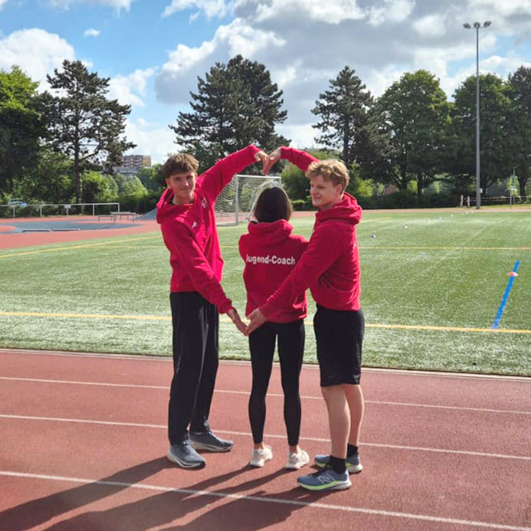 Auf dem Sportplatz formen die Jugend-Coaches Arvid Meier und Felix Gerke mit ihren Armen ein Herz – mitten darin steht Elly Miethke. Gemeinsam sind sie engagierte Gesichter des Projekts ‚Werde Jugend-Coach!‘ und zeigen: Herz und Teamgeist gehören hier zusammen.