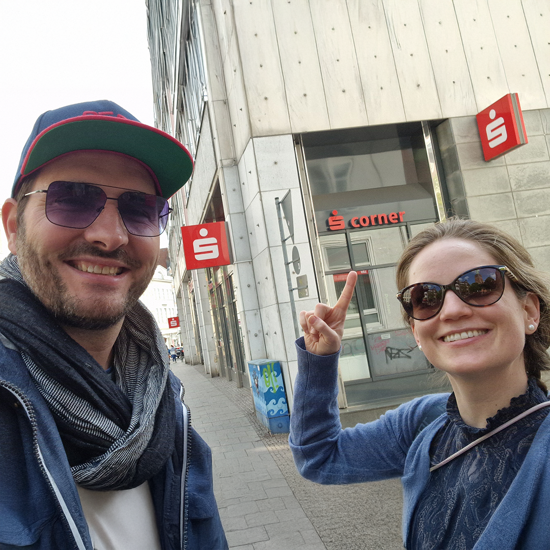 Florian und Dorothea von PEPE berlin stehen vor dem Sparkassen Corner – mit einem Lächeln deutet Doro auf die großen Buchstaben am Gebäude – ein Moment mit Augenzwinkern.