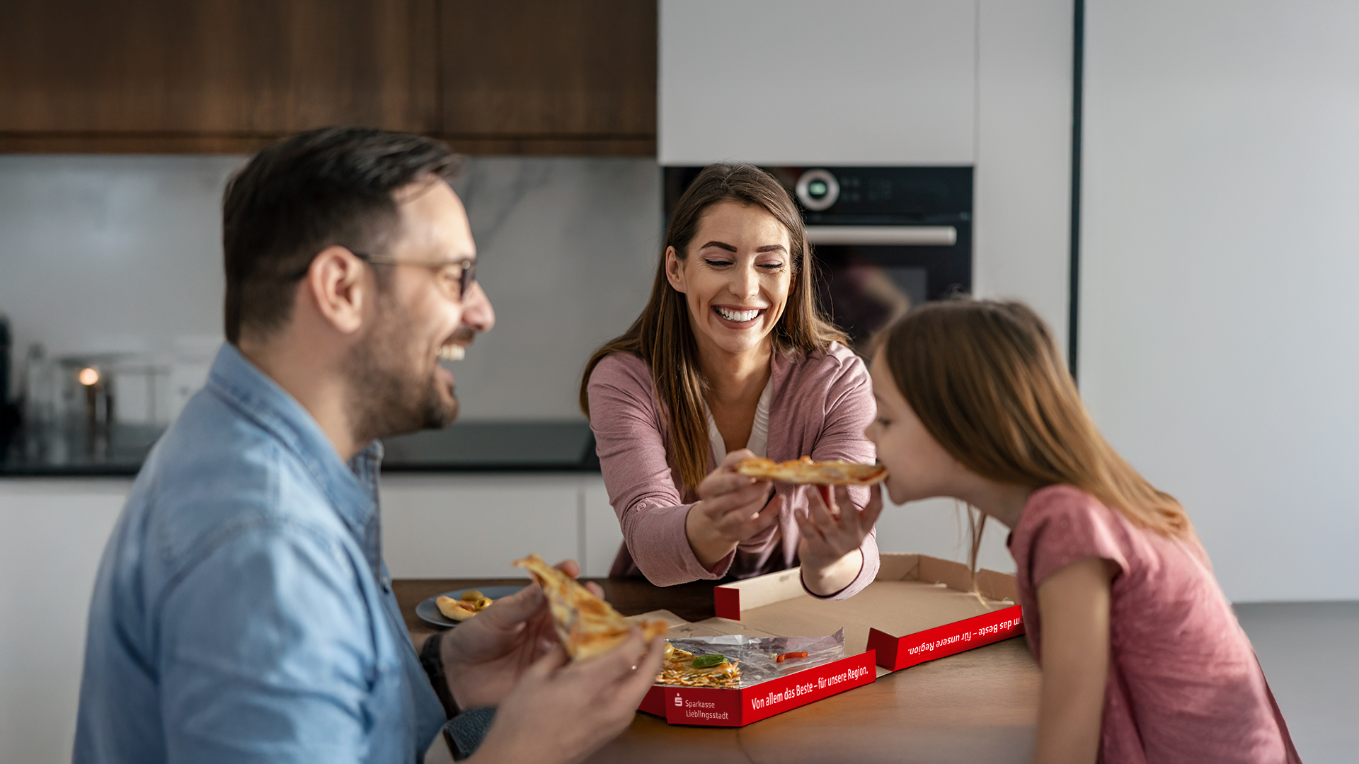Eine junge Familie (Mutter, Vater, Kind) sitzt Zuhause am Küchentisch und isst vergnügt zusammen Pizza.