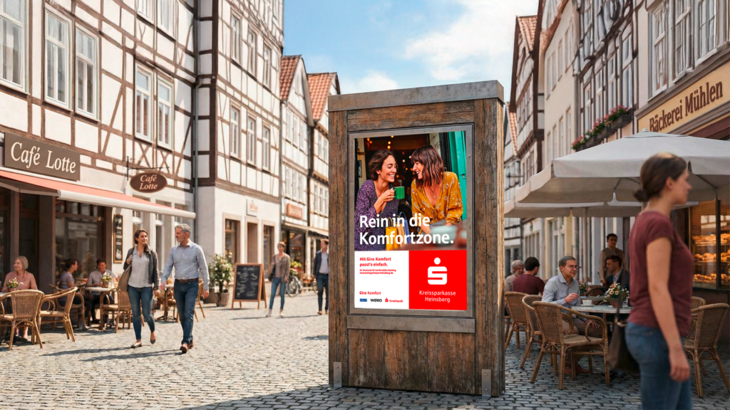 In einer Fußgängerzone steht zwischen vielen kleinen Cafes eine Plakatwand mit dem Motiv der KSK Heinsberg und der Aufschrift: Rein in die Komfortzone